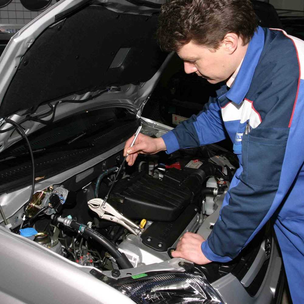 Mann in Arbeitskleidung beugt sich über geöffneten Motorraum und hält einen Stab mit Klemme über den Motorraum